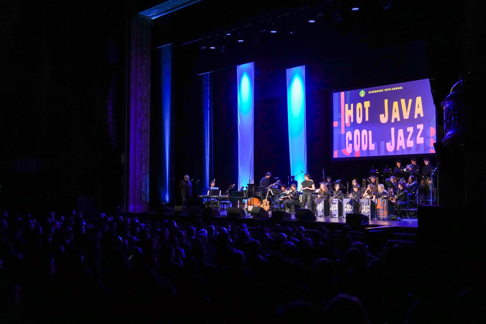 A jazz band performs on stage with blue lighting, while a large screen displays "Hot Java Cool Jazz." An audience is visible in the foreground.