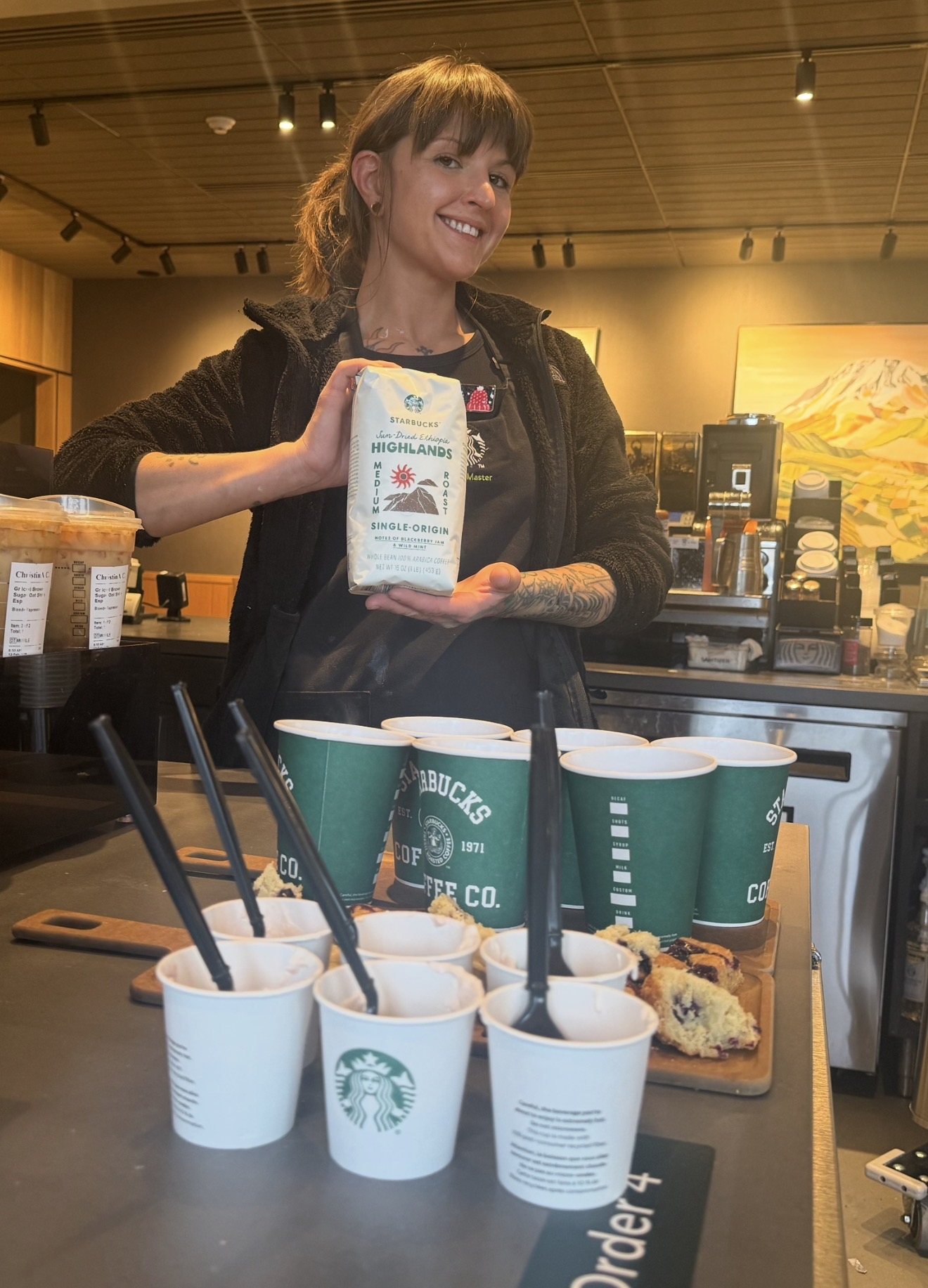 A barista holds a bag of Starbucks Highlands coffee in a café. Several cups with straws and a slice of baked goods are on the counter.