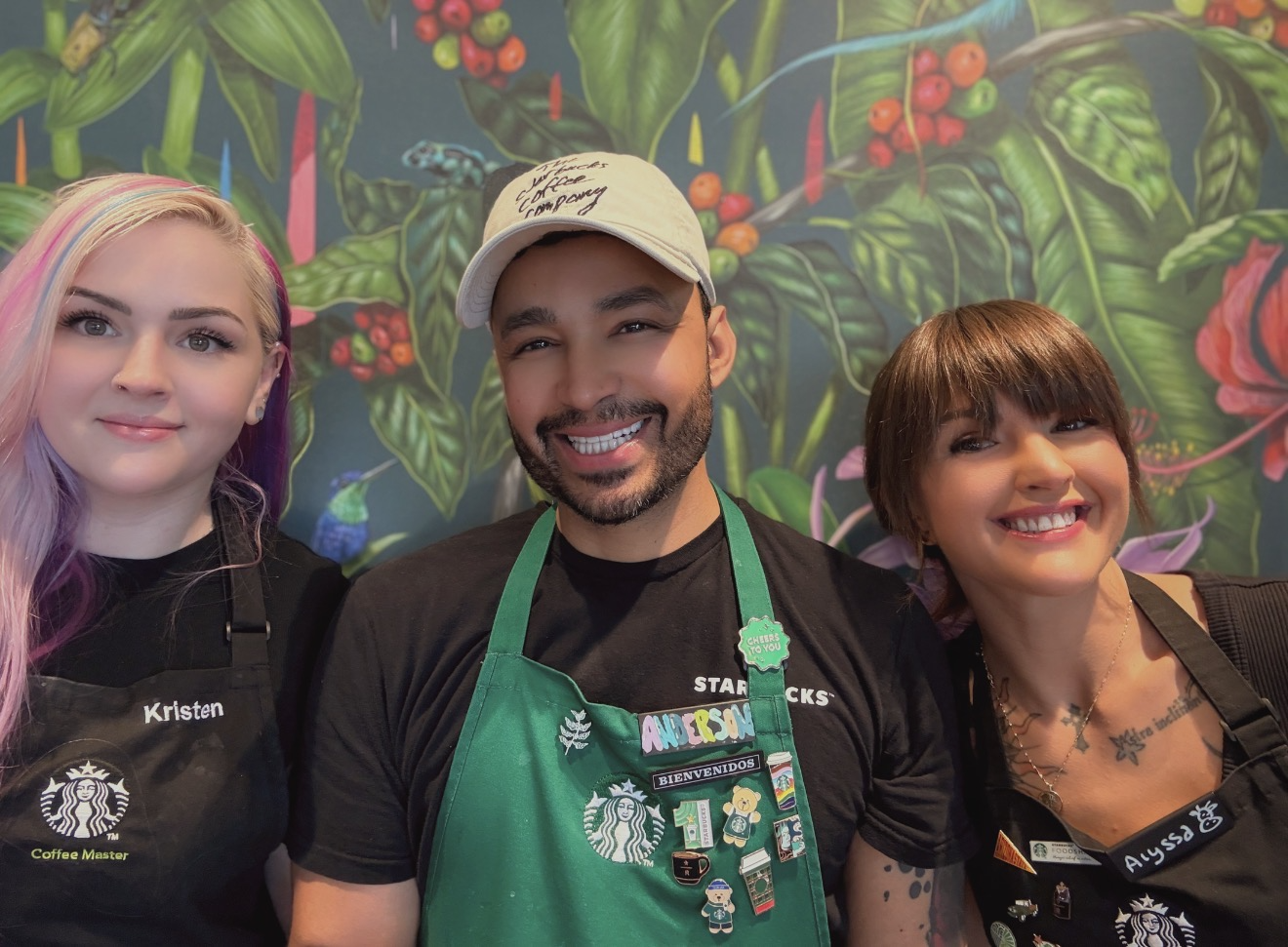 Three baristas smile in front of a vibrant, tropical-themed mural. They wear black aprons with Starbucks logos, showcasing various pins.