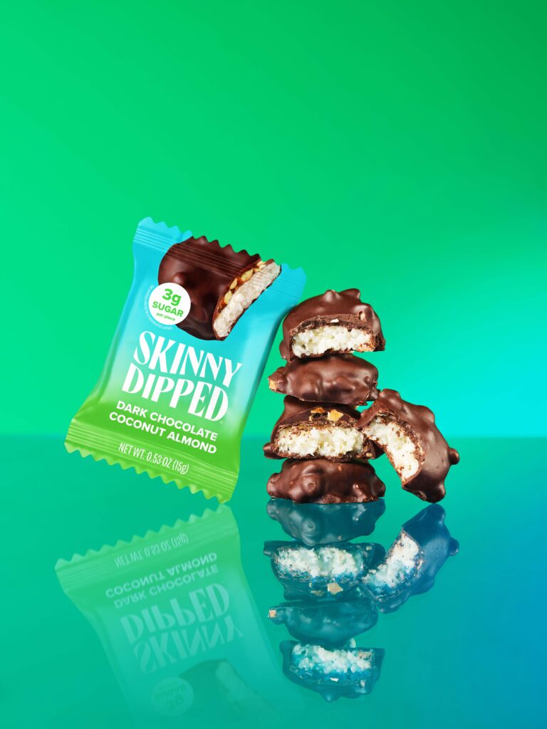 A vibrant green background showcases a package of Skinny Dipped Dark Chocolate Coconut Almond snacks, with several pieces stacked beside it. The snacks are coated in dark chocolate, revealing a coconut filling inside.