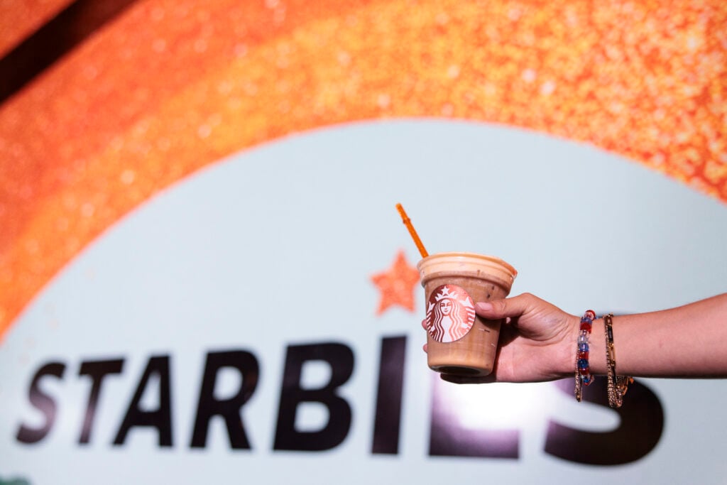 A hand holds a Starbucks cup filled with a blended drink, featuring a bright orange straw. The background displays a colorful, sparkly design with the word "Starbies.