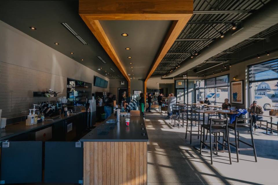 A modern café interior features a long counter with coffee equipment, wooden accents, and ample seating. Large windows allow natural light, highlighting patrons enjoying their time.