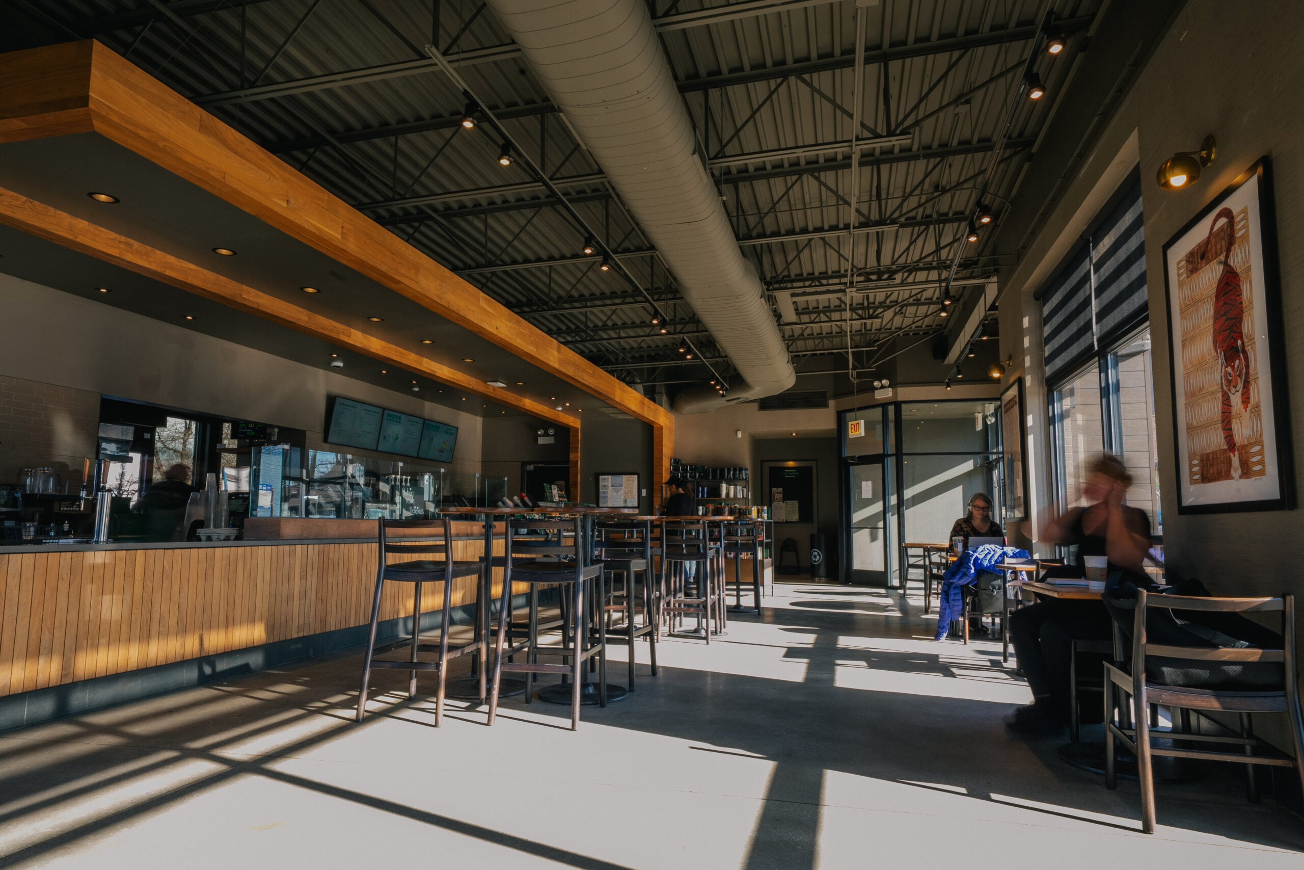 A modern café interior featuring wooden accents, high tables, and chairs. Large windows allow natural light to create shadows on the floor.