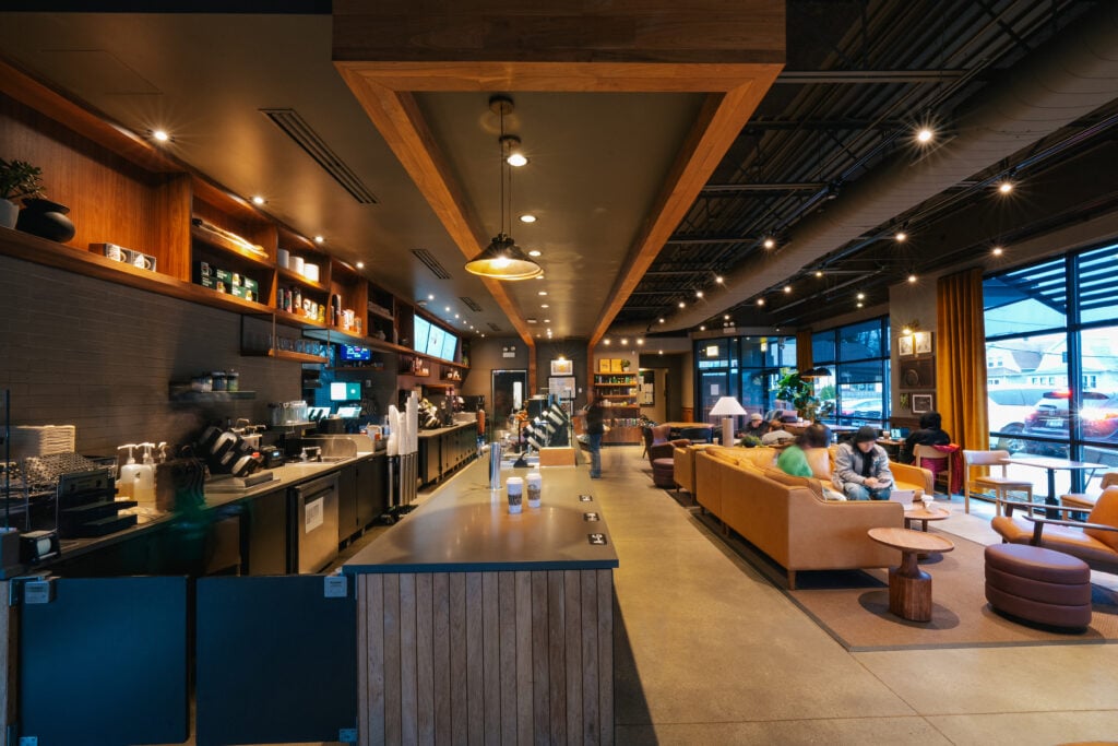 A modern coffee shop interior featuring a long counter with coffee machines, seating areas with couches, and warm lighting.
