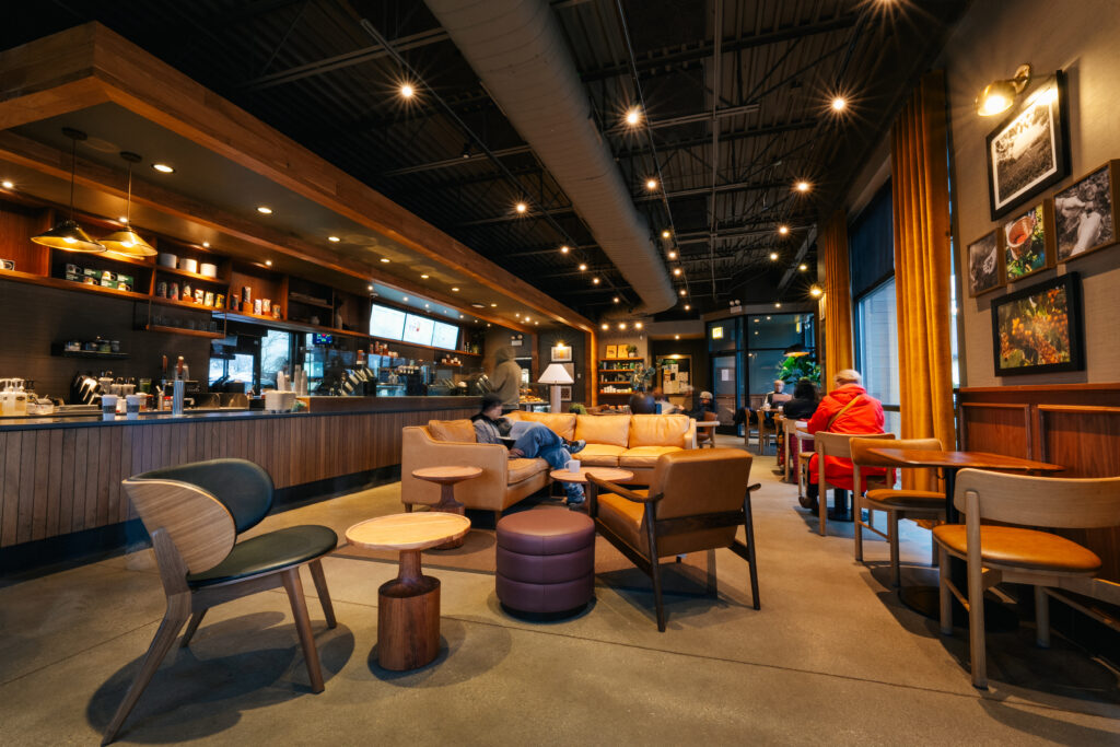 A cozy coffee shop interior featuring warm lighting, wooden accents, and comfortable seating. Patrons are scattered throughout, some relaxing on a leather couch, while others occupy tables. A counter with coffee equipment and menu displays is visible in the background.