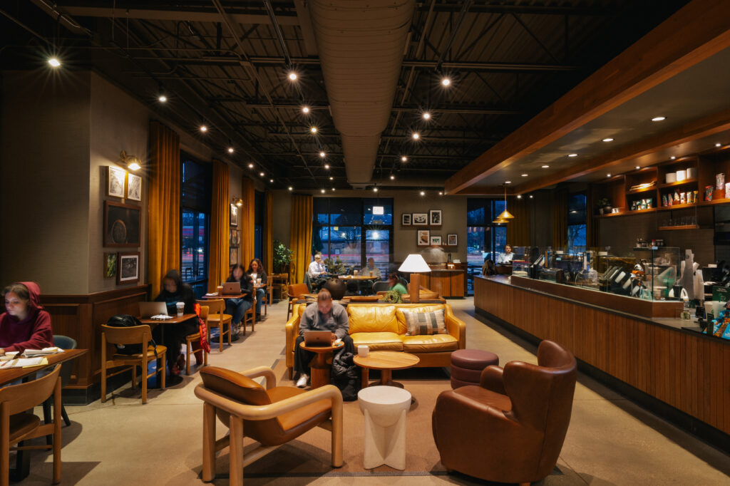 A cozy café interior featuring a mix of seating options, including a yellow leather sofa and wooden chairs. Soft lighting and large windows create a warm atmosphere. People are working on laptops, and a counter with a barista is visible in the background.