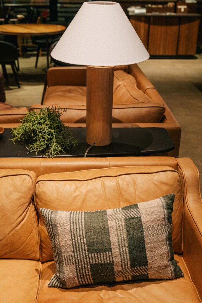 A cozy leather sofa features a decorative pillow with a geometric pattern in green and beige. A wooden lamp with a white shade and a small plant sit nearby.