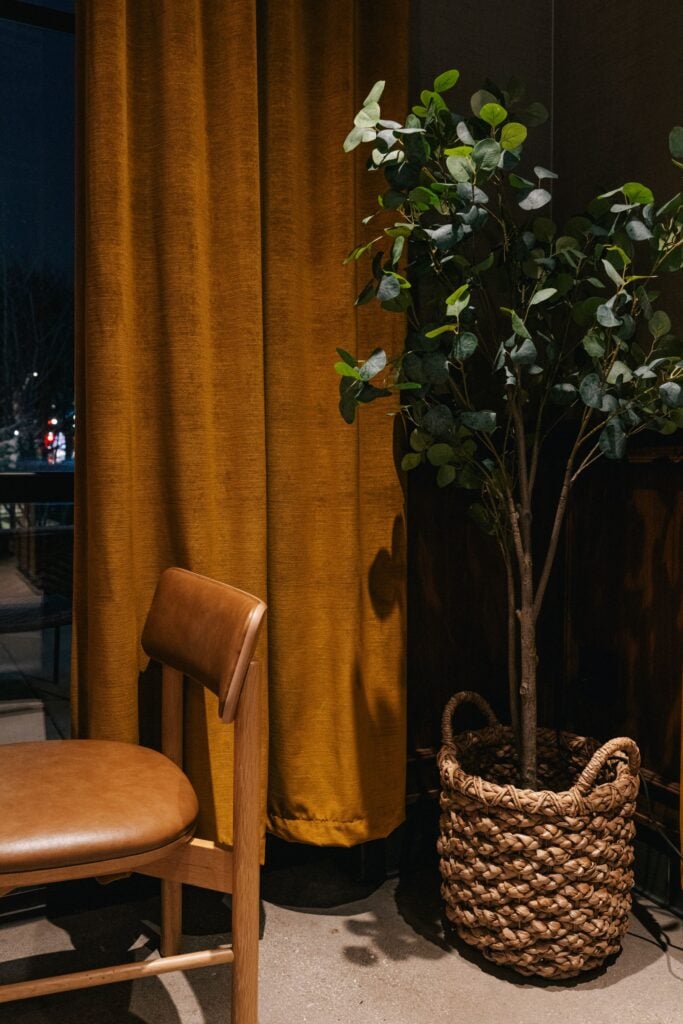 A wooden chair with a brown seat is positioned next to a tall potted plant in a woven basket. Behind them, there are mustard-colored curtains.