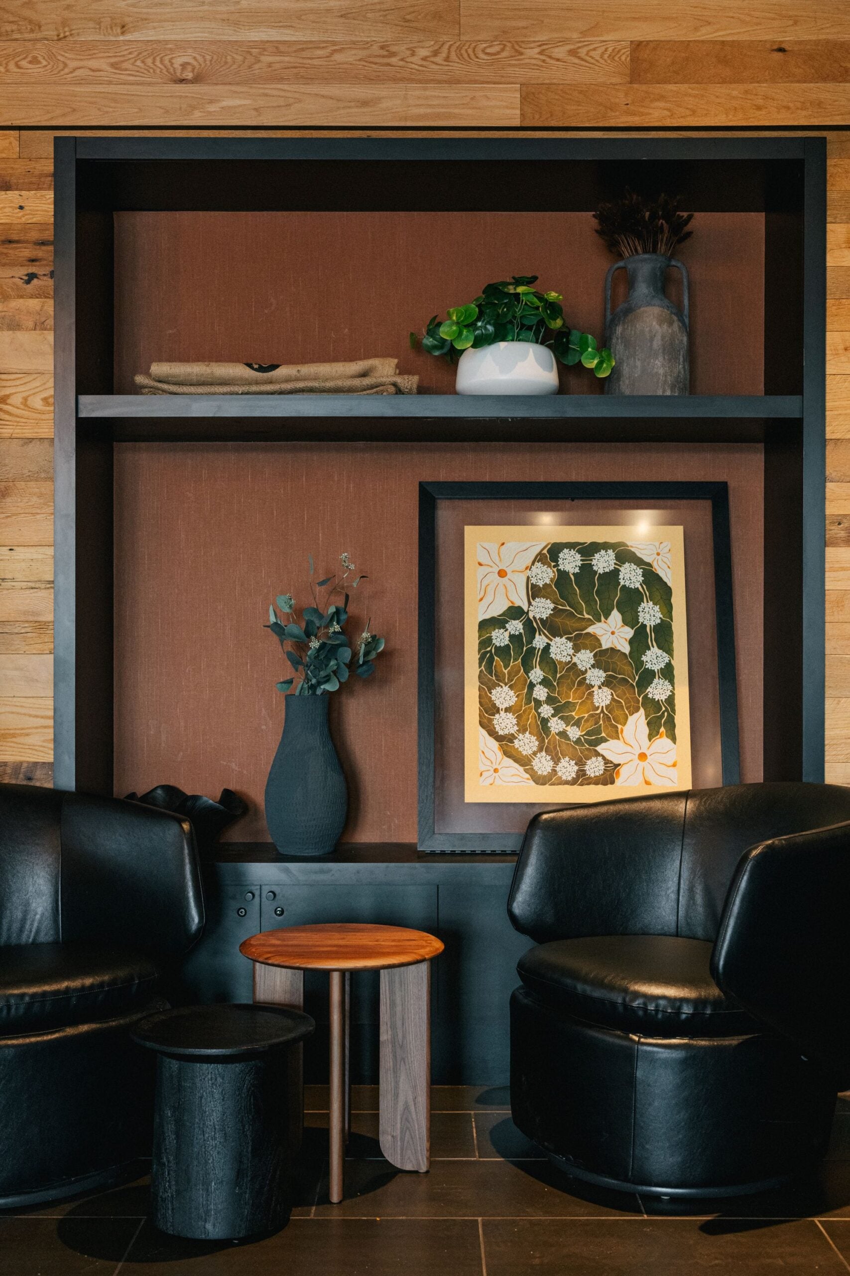 A cozy corner features two black leather chairs, a wooden side table, and a black stool. A shelf above displays a vase, a potted plant, and framed artwork.