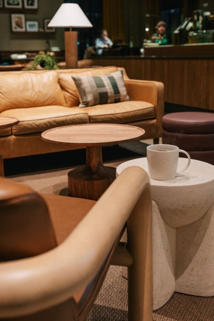 A cozy seating area features a tan leather sofa with a plaid pillow, wooden tables, and a white mug on a textured stone table.