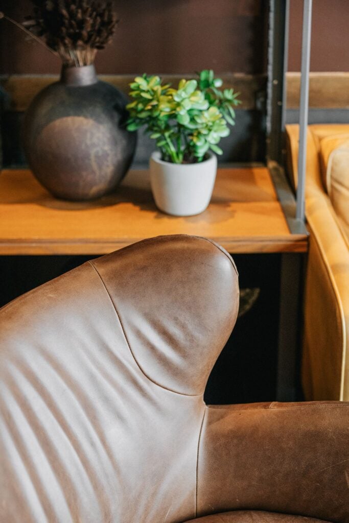 A close-up of a brown leather chair's armrest, with a decorative plant and a dark vase in the background on a wooden shelf.