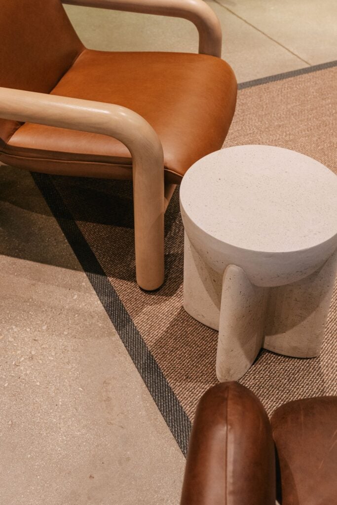 A modern lounge chair with a brown leather seat and light wooden arms is positioned next to a round, textured concrete side table. The setting features a woven rug and a smooth floor surface.