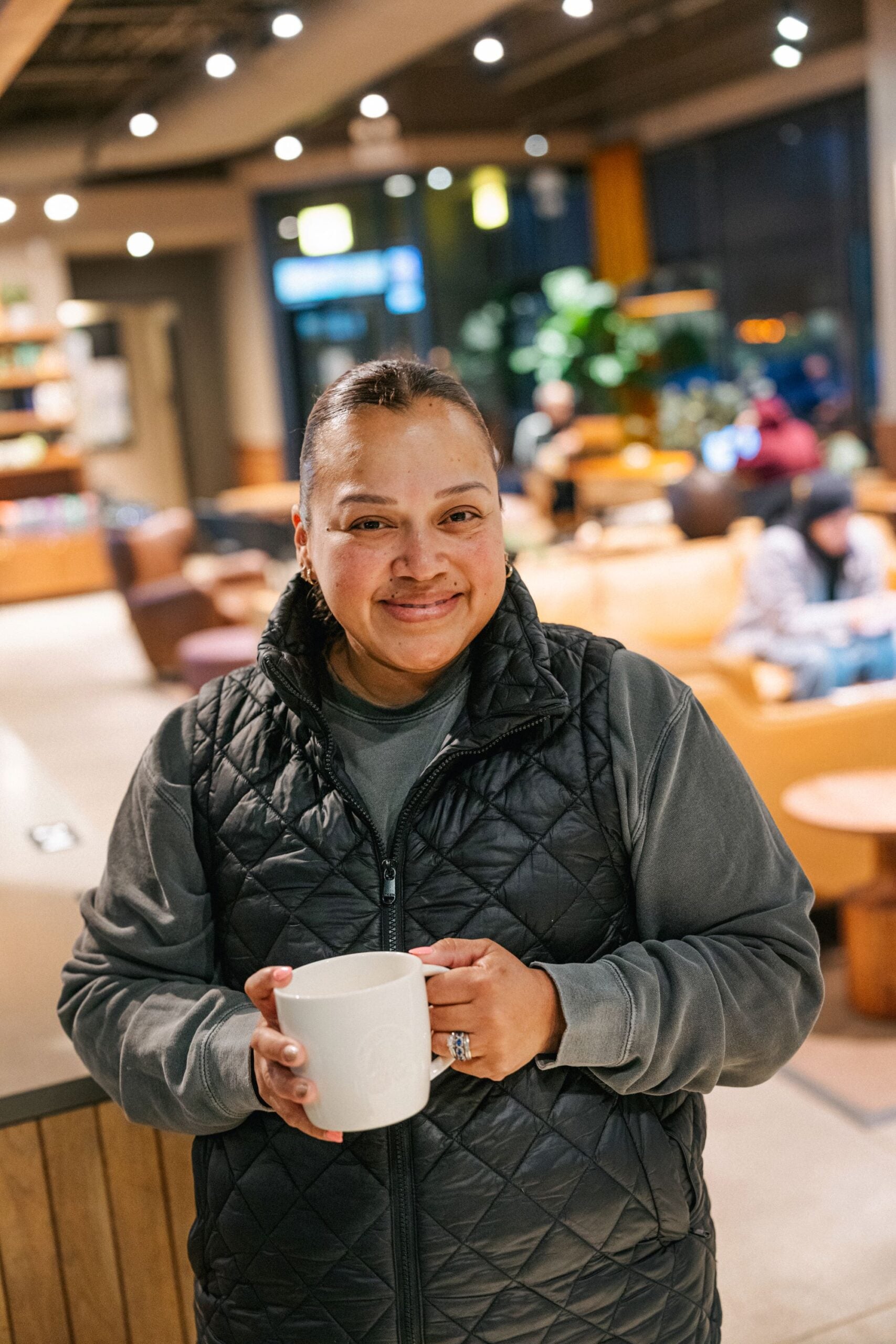 A woman in a black quilted vest holds a white mug, smiling warmly. The background features a cozy café setting with soft lighting and seating.