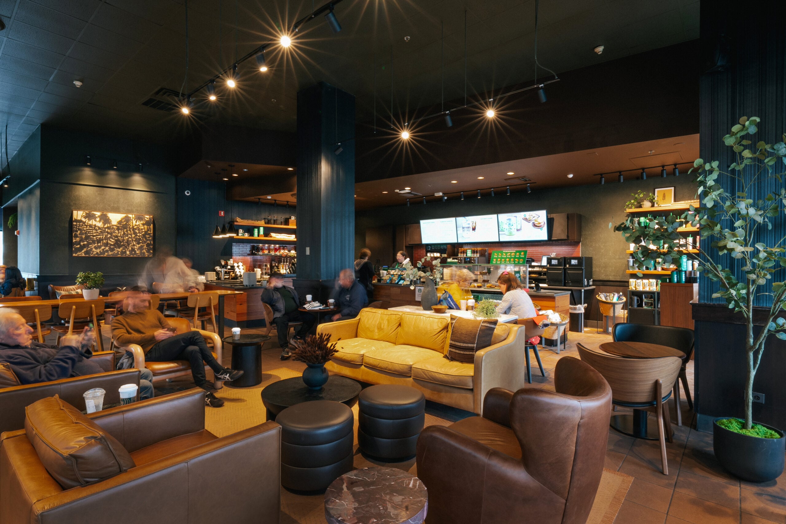 A cozy café interior featuring a yellow sofa, dark wood furniture, and a coffee counter with menu displays. Patrons are seated and mingling.