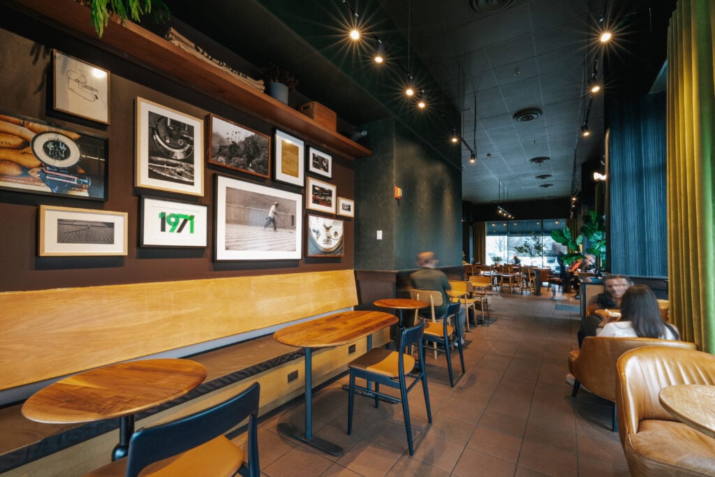 A cozy café interior features wooden benches and tables, framed artwork on dark walls, and warm lighting. Patrons are seated throughout.