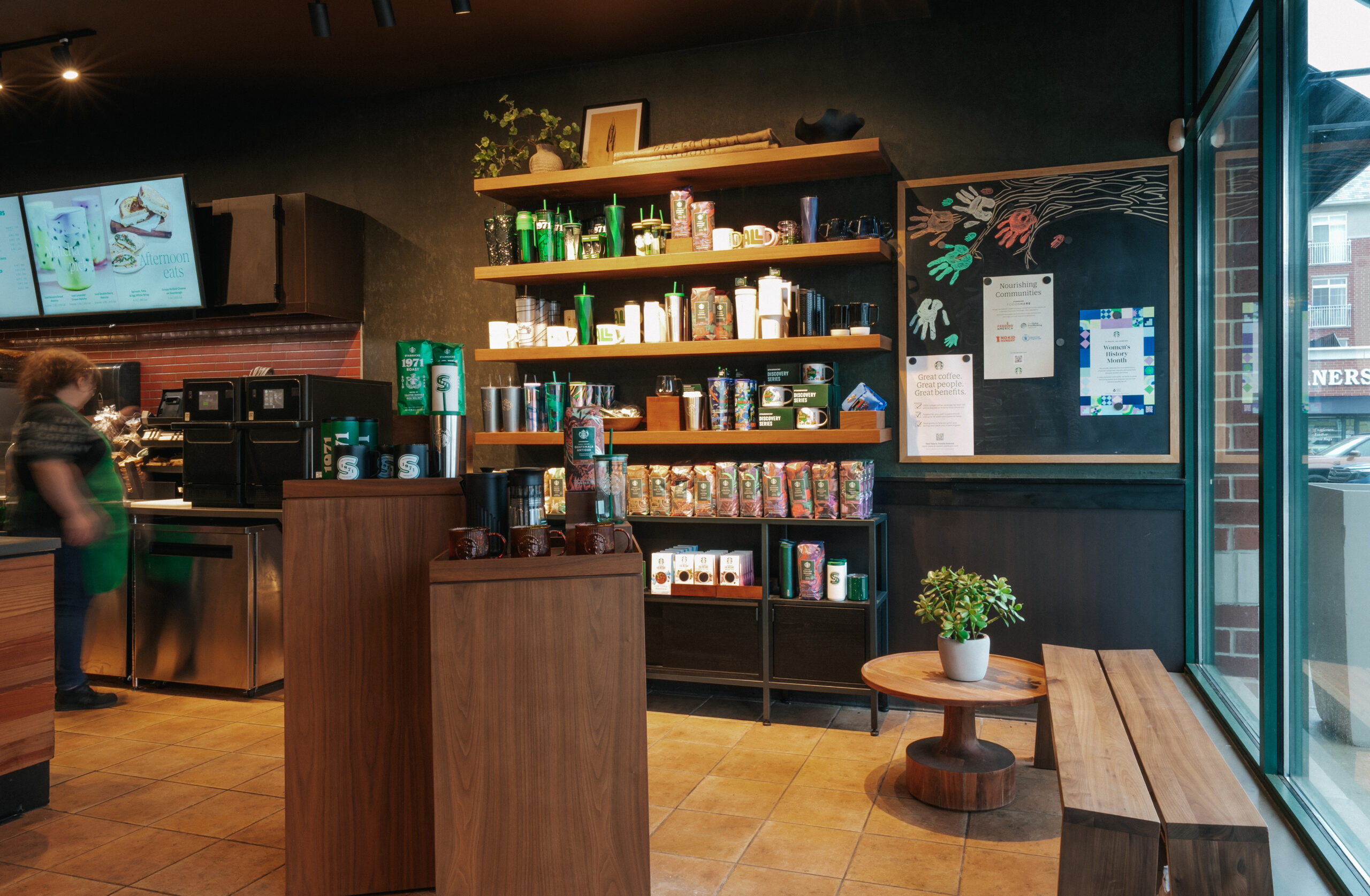 A cozy coffee shop interior featuring wooden shelves stocked with mugs, tumblers, and coffee bags. A small table and plant add warmth.