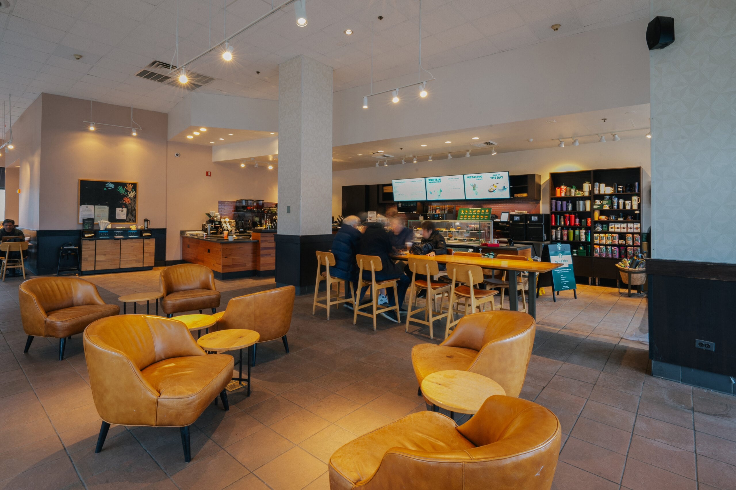 A cozy café interior featuring wooden furniture, including brown leather chairs and tables. A coffee counter is visible with menu screens and various drink options displayed. Customers are seated at tables, creating a relaxed atmosphere.