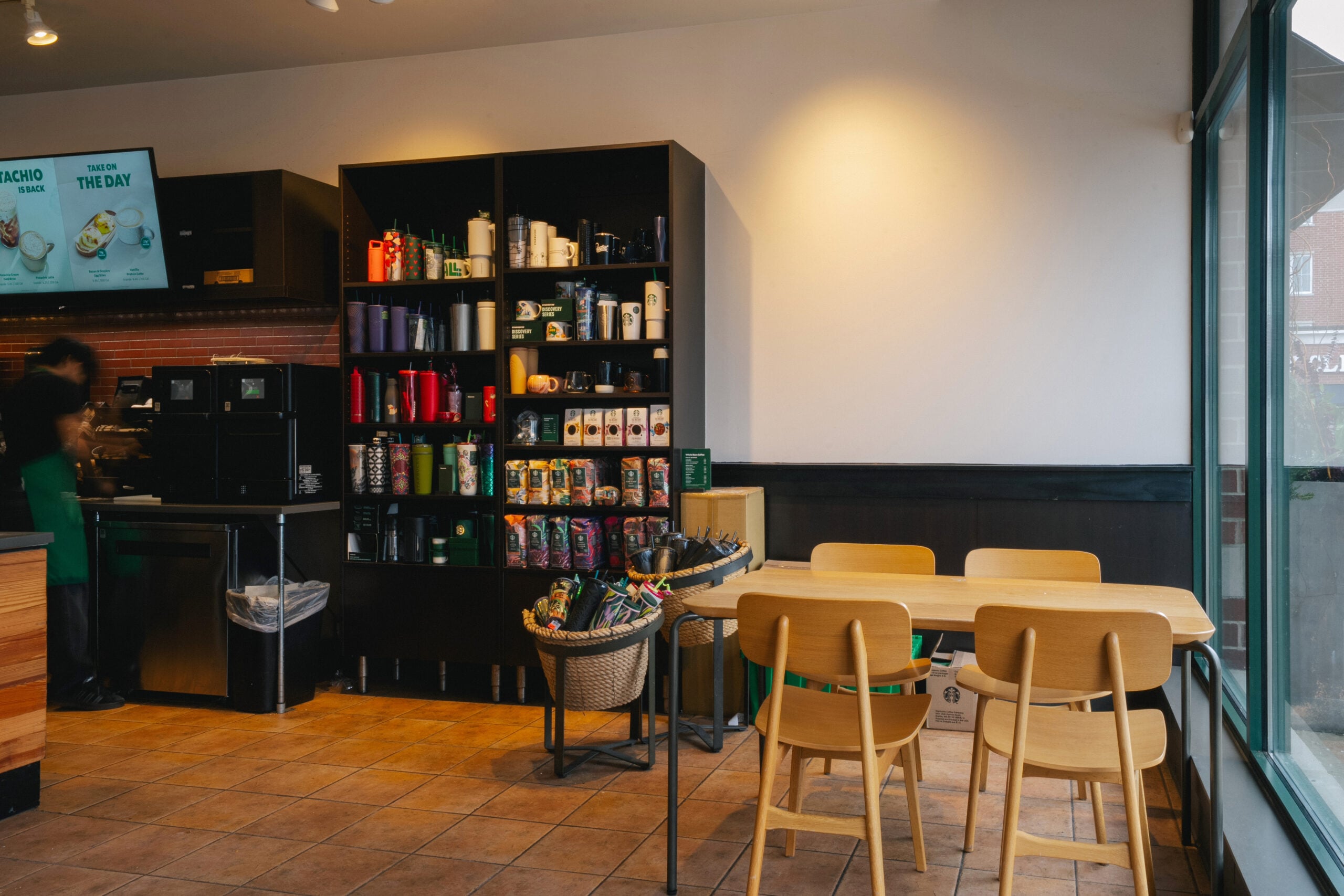 A cozy café interior featuring a black shelving unit filled with various drinkware and coffee products. There are wooden tables and chairs, with a couple of baskets holding items. The floor is tiled, and the walls are mostly bare.