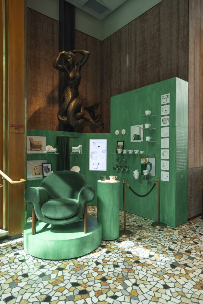 A vibrant green display features a plush armchair on a circular pedestal, surrounded by shelves showcasing various ceramic items. A bronze mermaid statue is positioned above, and the floor is decorated with a colorful mosaic pattern.