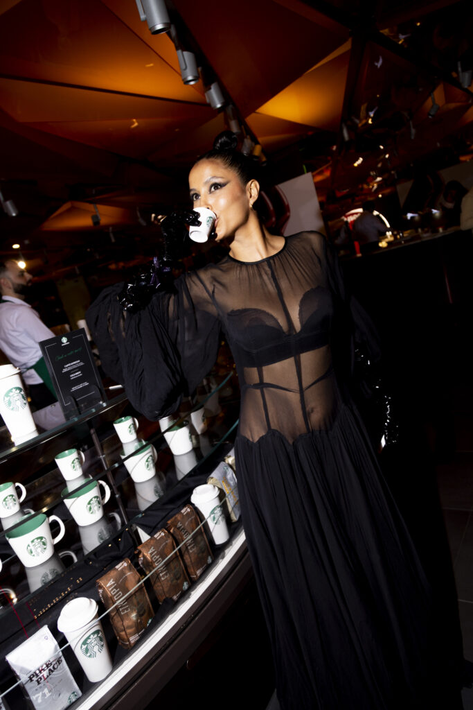 A woman in a sheer black dress poses with a coffee cup in a café setting. Behind her, there are Starbucks mugs and coffee bags on display. The ambiance features warm lighting and modern decor.
