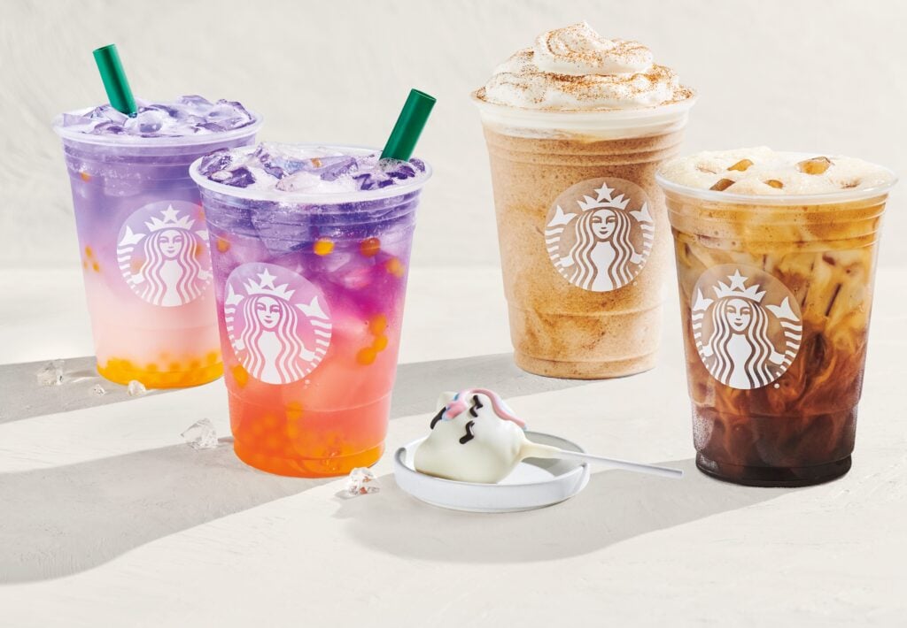 Three colorful drinks with ice, featuring a purple and pink beverage with tapioca pearls, a creamy coffee drink, and an iced coffee. A small dessert shaped like a unicorn is on a plate beside them.