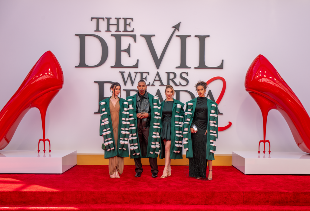 Four individuals pose on a red carpet in front of a large sign reading "THE DEVIL WEARS PRADA 2." They wear stylish green coats with white accents. Two oversized red high heels are displayed prominently on either side. The setting is vibrant and fashion-forward.