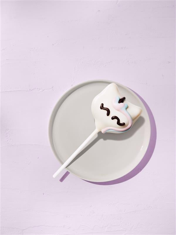 A decorated cake pop shaped like a cat's face, featuring white icing, pastel accents, and chocolate detailing, sits on a white plate.