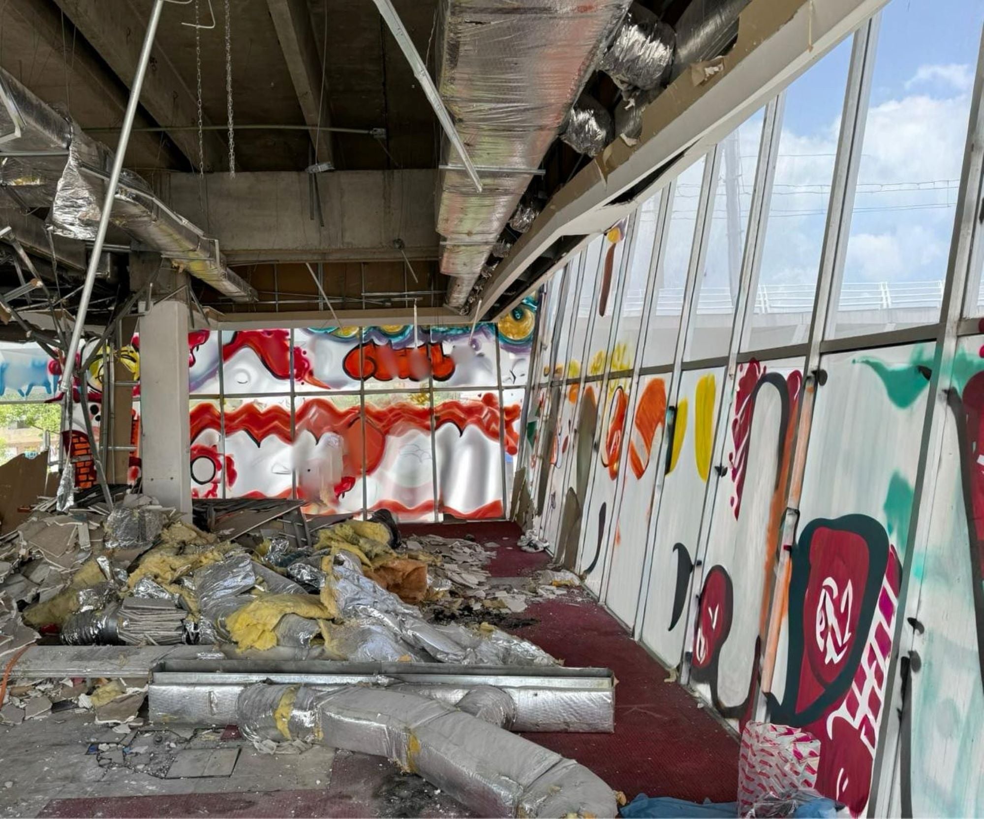 An interior space shows debris scattered on the floor, including insulation and metal ductwork. Colorful graffiti decorates the walls, contrasting with the exposed structural elements and unfinished appearance. Natural light filters through large windows, revealing a partially demolished environment.
