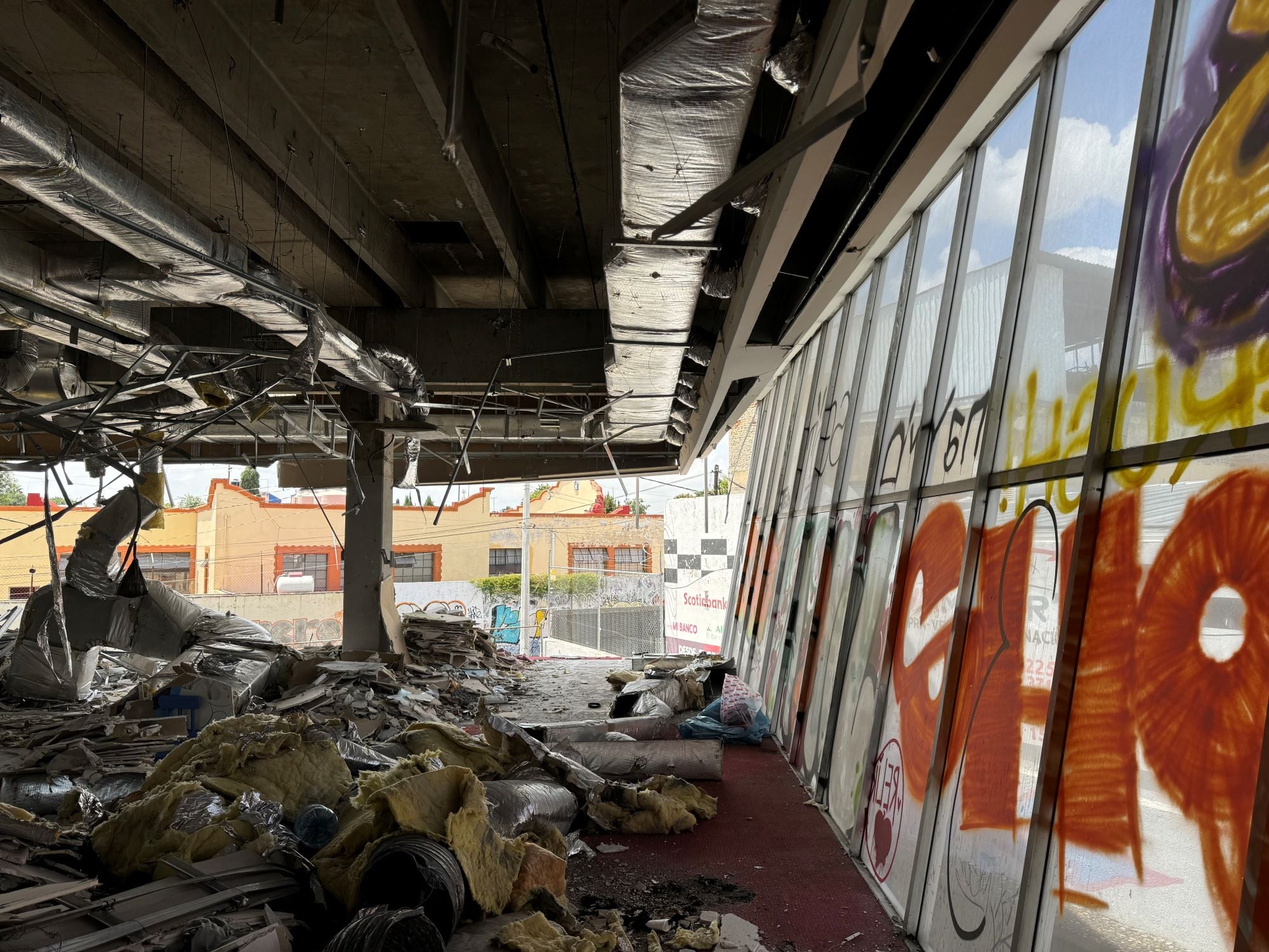 An abandoned interior space with debris scattered on the floor, exposed insulation, and graffiti on large glass windows.