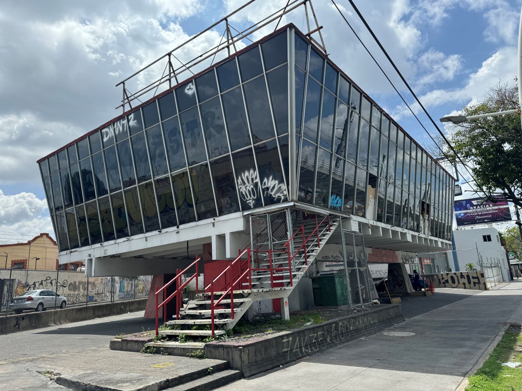 A modern, glass-fronted building with a slanted roof, elevated on stilts. The exterior features graffiti and red stairs leading up.