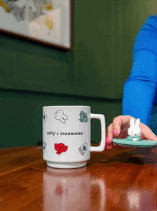 A white mug featuring Miffy and Starbucks branding, decorated with colorful illustrations. A hand holds a small green plate with a Miffy figurine.
