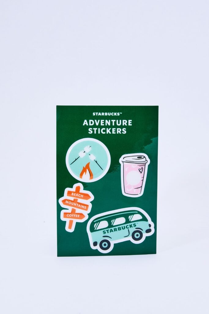 A green sticker sheet featuring Starbucks-themed adventure stickers, including a campfire with marshmallows, a coffee cup, and a directional sign with "Beach," "Mountains," and "Coffee.