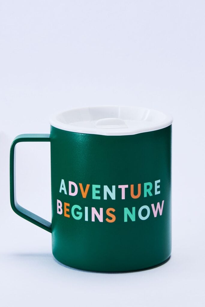 A green mug with a white lid features the phrase "ADVENTURE BEGINS NOW" in colorful letters.