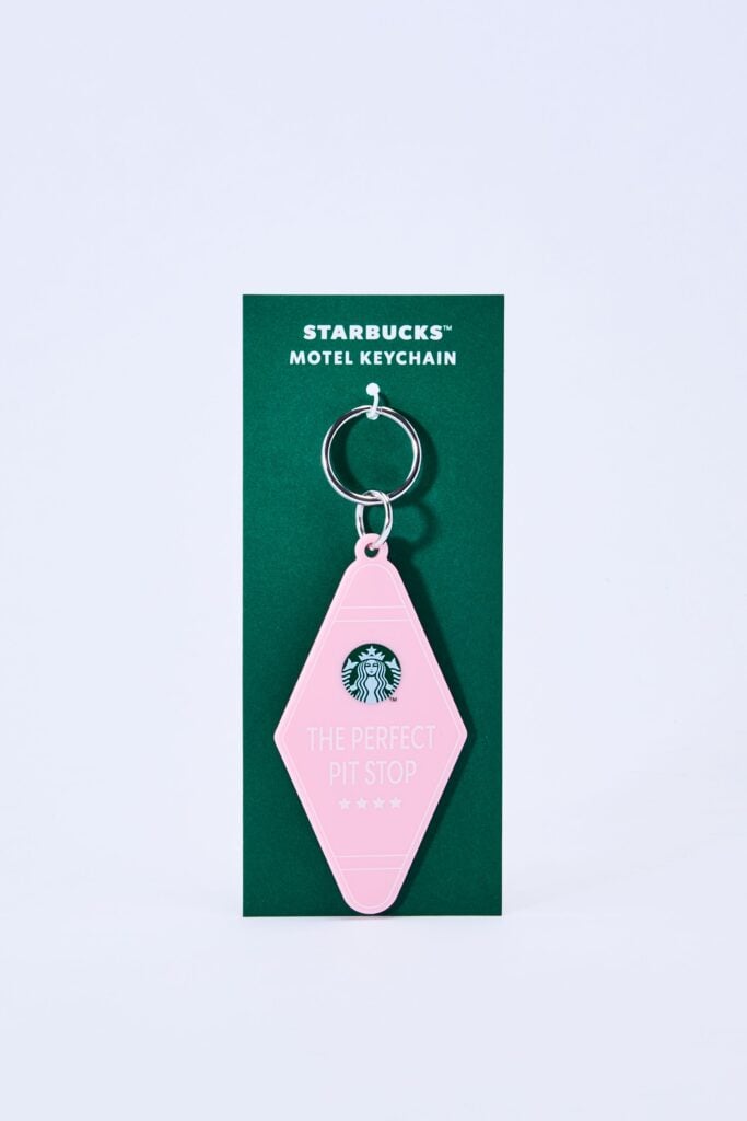 A pink motel keychain shaped like a diamond, featuring the text "THE PERFECT PIT STOP" and a Starbucks logo, attached to a silver keyring. The keychain is displayed on a green card labeled "STARBUCKS™ MOTEL KEYCHAIN.