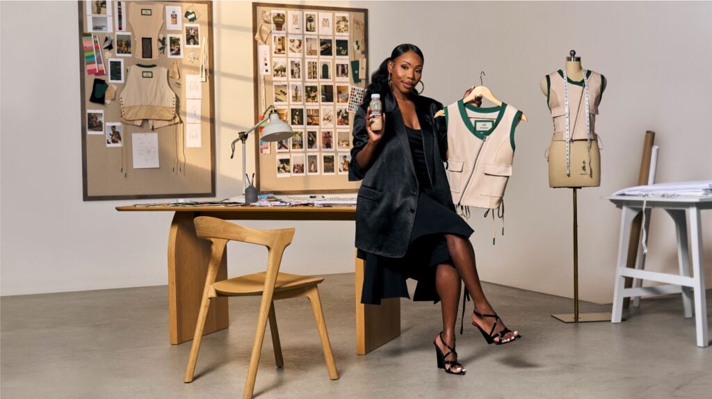 A woman in a black outfit holds a Starbucks drink in one hand and a beige vest on a hanger in the other. She stands beside a wooden table with design materials and a lamp, against a backdrop of pinned photos and sketches.
