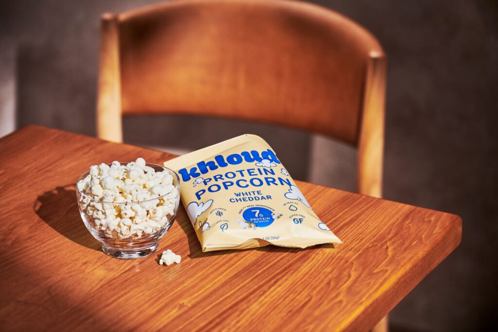 A clear bowl filled with popcorn sits on a wooden table next to a bag of Khloud Protein Popcorn in White Cheddar flavor.