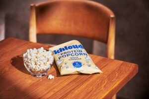 A clear bowl filled with popcorn sits on a wooden table next to a bag of Khloud Protein Popcorn in White Cheddar flavor.
