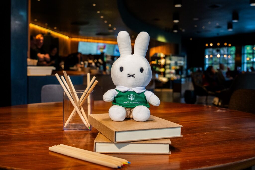 A plush bunny toy wearing a green shirt sits on two stacked books. Nearby, a glass container holds wooden sticks, and colored pencils are scattered on the table. The background features a café setting with blurred patrons.