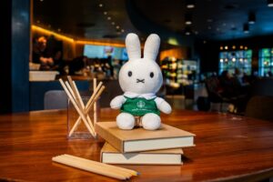 A plush bunny toy wearing a green shirt sits on two stacked books. Nearby, a glass container holds wooden sticks, and colored pencils are scattered on the table. The background features a café setting with blurred patrons.