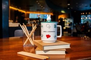 A decorative mug featuring Miffy and Starbucks branding sits atop two stacked notebooks. Nearby, a glass holds wooden sticks.