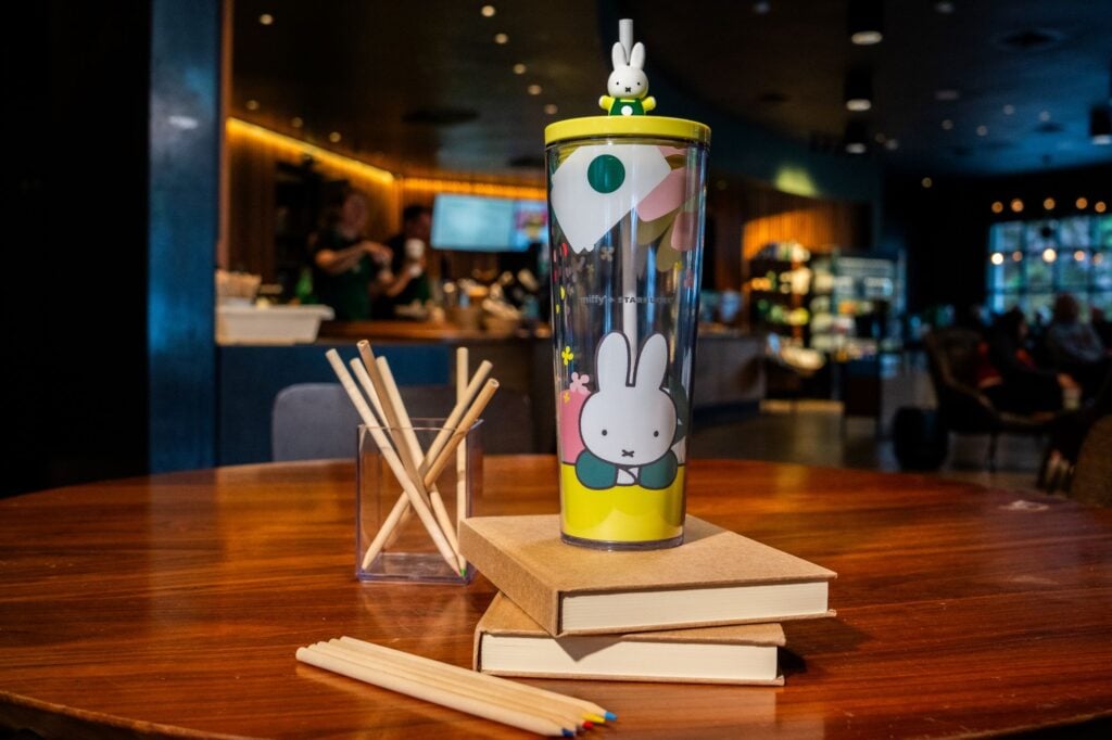 A colorful tumbler featuring a cartoon bunny sits on a wooden table, surrounded by stacked books and a glass holder with wooden straws.