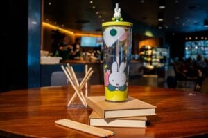 A colorful tumbler featuring a cartoon bunny sits on a wooden table, surrounded by stacked books and a glass holder with wooden straws.
