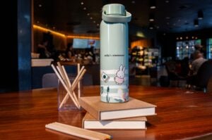 A mint green thermos featuring a Miffy character design sits on a wooden table, alongside stacked notebooks and a container of wooden pencils. The background shows a cozy café atmosphere.