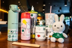 A collection of colorful mugs and tumblers featuring Miffy designs, alongside a plush Miffy toy. The items are arranged on a wooden table.