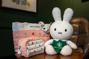 A plush Miffy toy wearing a green Starbucks outfit sits next to a pink and white tote bag adorned with cute designs and a small matching pouch.