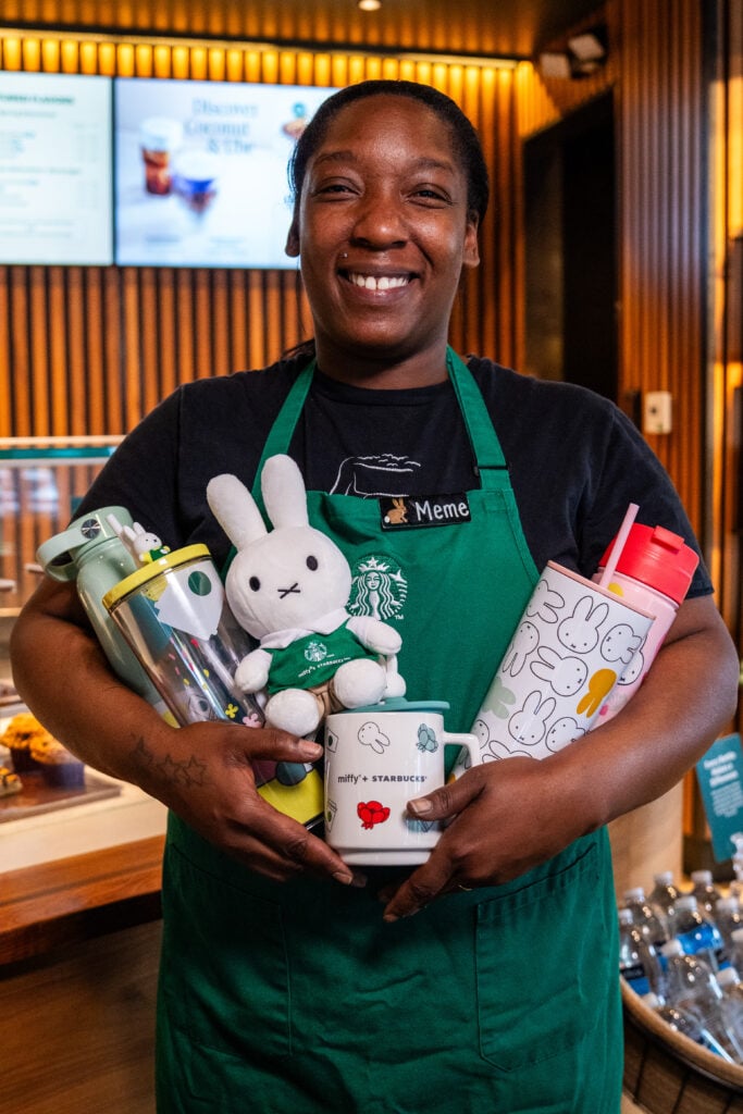 A smiling barista in a green apron holds various drinkware, including a plush bunny, colorful tumblers, and a mug featuring a character design. The background shows a café setting with a menu display.