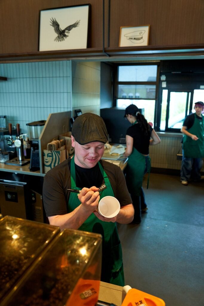 A barista in a green apron writes on a white cup while smiling. The café has a modern interior with other staff members in the background.