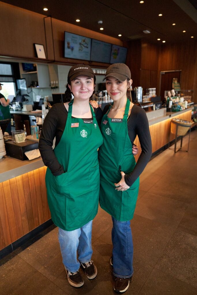 Two baristas in green aprons pose together in a Starbucks store. The background features a coffee counter and menu screens.
