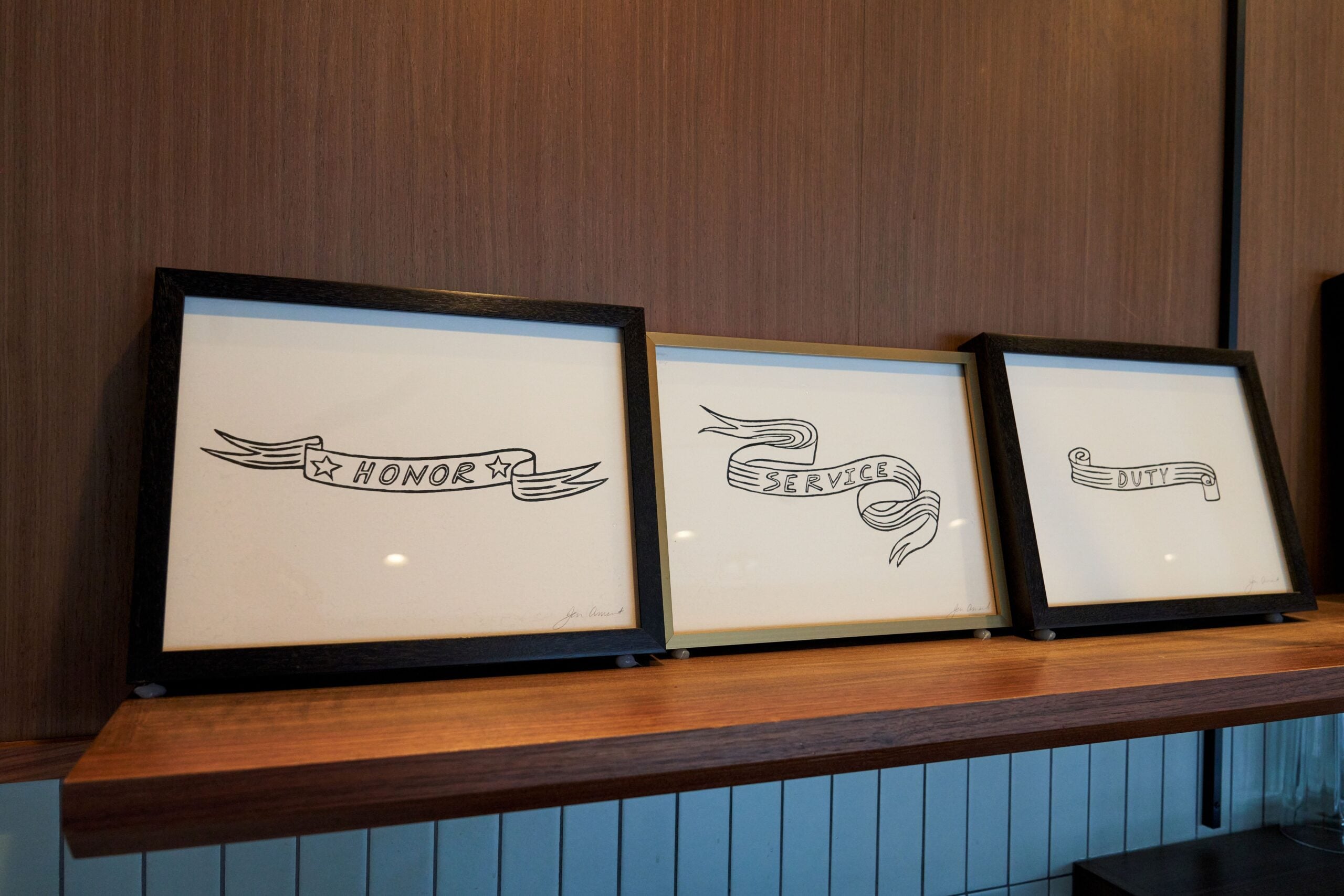 Three framed illustrations on a wooden shelf feature decorative ribbons with the words "HONOR" and "SERVICE" in bold lettering. The background is a textured wood panel, and the shelf is complemented by a blue tiled base.