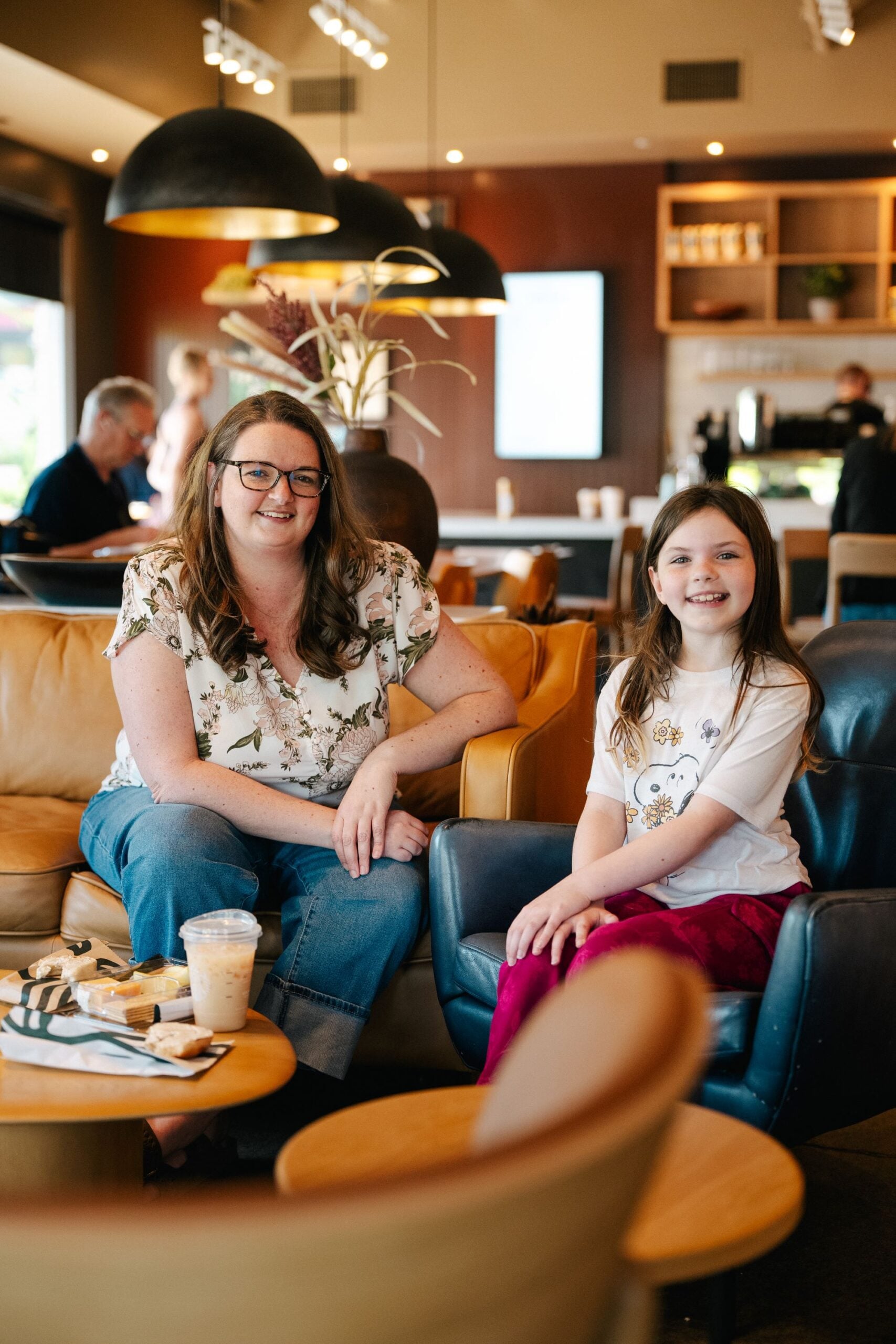 A woman and a girl sit together in a cozy café, smiling. The woman wears a floral top and jeans, while the girl is in a white shirt and red pants. A table in front holds snacks and drinks. The café has a warm, inviting atmosphere with modern decor.